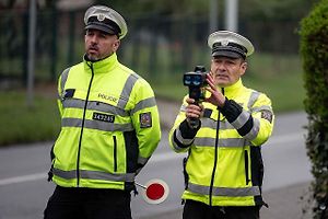 Policista v uniformě obsluhuje ruční laserový měřič rychlosti, jehož signál se zakázané aktivní antilasery snaží neúspěšně rušit.